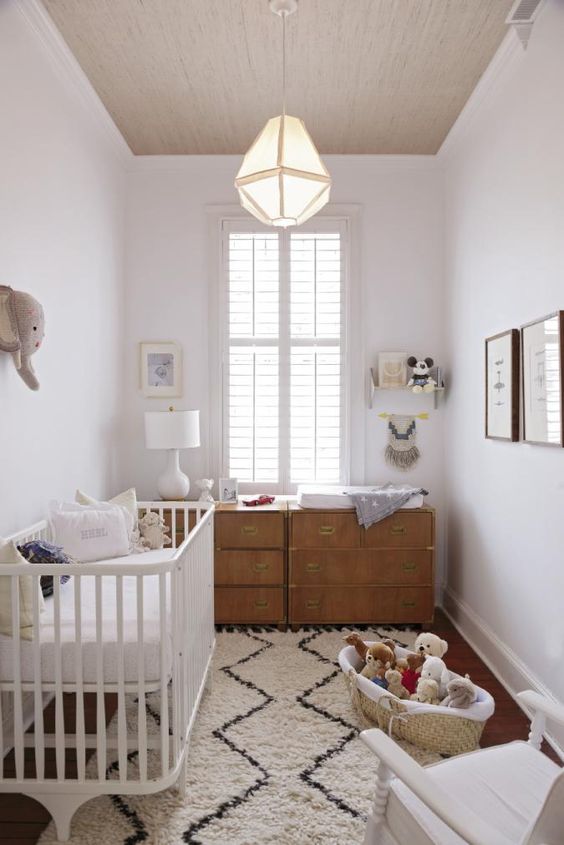 a neutral nursery with a double dresser, a white rocker and crib, a pendant lamp, a basket with toys and a gallery wall