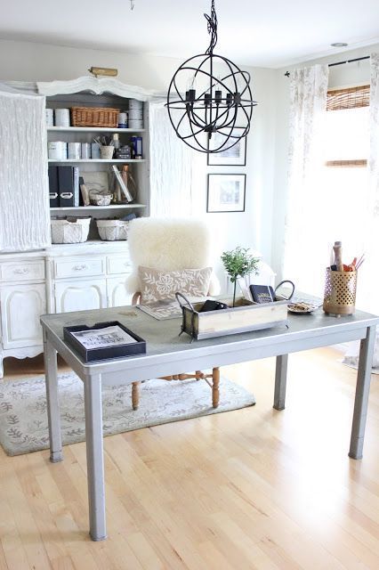 a neutral rustic home office with a large vintage white buffet, a light grey desk, a white chair, a metal sphere chandelier