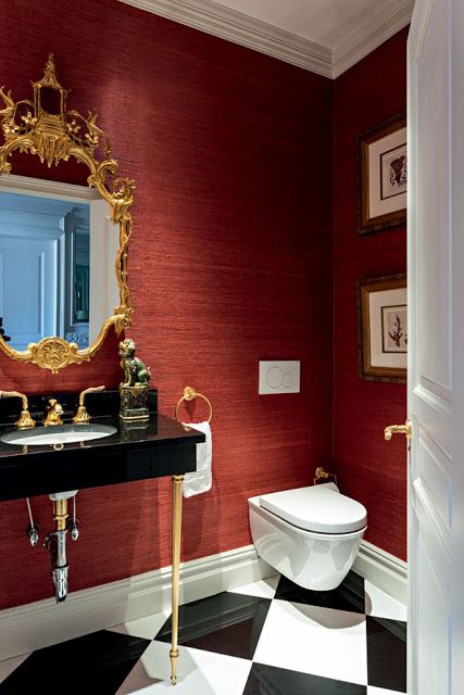 a refined and chic red bathroom with black and white tiles on the floor, a black and gold vanity, a gold mirror and some artworks