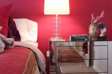 a refined mid-century modern bedroom with a red accent wall, bold red and burgundy bedding, a printed rug and a mirror nightstand with a chic lamp