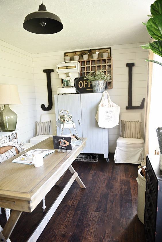 a relaxed neutral rustic home office with a white storage cabinet, a light-colored wooden desk, vintage lamps and greenery in buckets