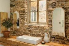 a rustic bathroom with a stone accent wall, a wood slab console table, arched mirrors and a plant in a pot