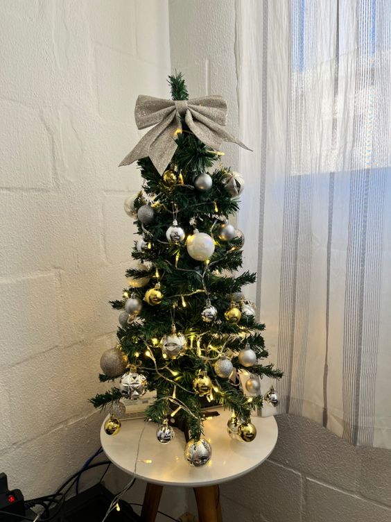 a small and pretty tabletop Christmas tree with lights and gold and silver ornaments plus a silver glitter bow on top