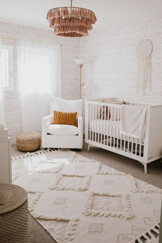 a sweet neutral nursery with wallpaper walls, white furniture, layered rugs, a pink tassel chandelier and some pillows