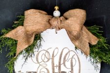 a vintage rustic Christmas ornament of a wooden plaque with calligraphy, a burlap bow and greenery on top