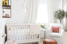 a welcoming neutral nursery with printed wallpaper, white furniture, a tassel chandelier, a leather ottoman and a woven basket plus greenery