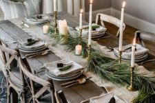 a Nordic Christmas tablescape with a white burlap runner, fir branches, candles, grey plates, bells and an upcovered wooden table and chairs