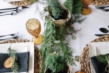a Scandinavian Christmas tablescape with woven chargers, colored glasses, a greenery and foliage runner, black napkins and dried citrus