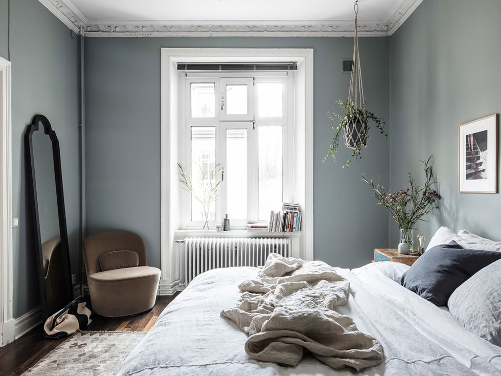 a Scandinavian bedroom with character, grey walls, stucco on the ceiling, a mirror in a black frame, a tan chair and some potted plants