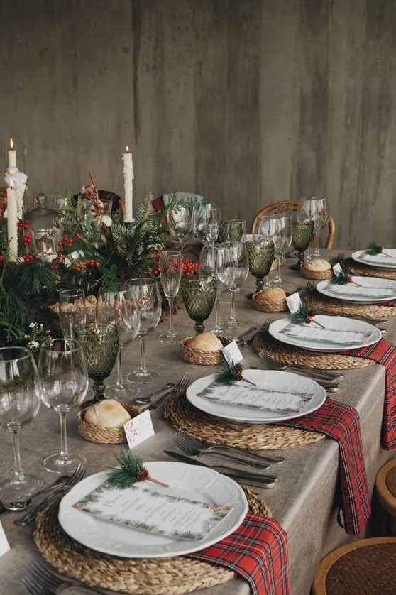 a beautiful Christmas tablescape with evergreens, berries, tall candles, green glasses, woven placemats, red napkins and printed menus