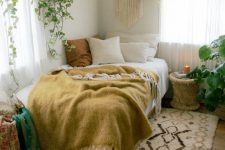 a boho bedroom with a bed and mustard bedding, a printed rug, potted plants, a rattan table and a macrame piece