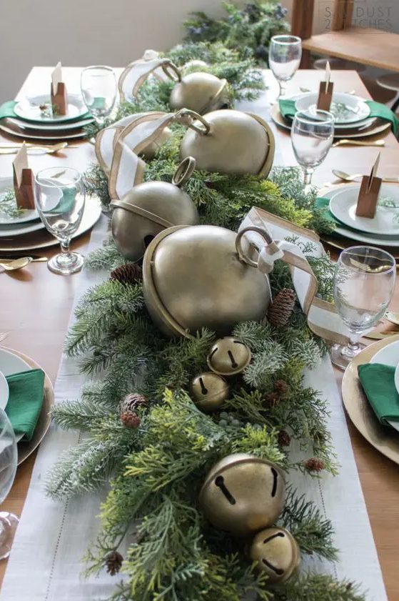 a bold Christmas tablescape with an evergreen runner with pinecones and oversized bells, metallic placemats and emerald napkins