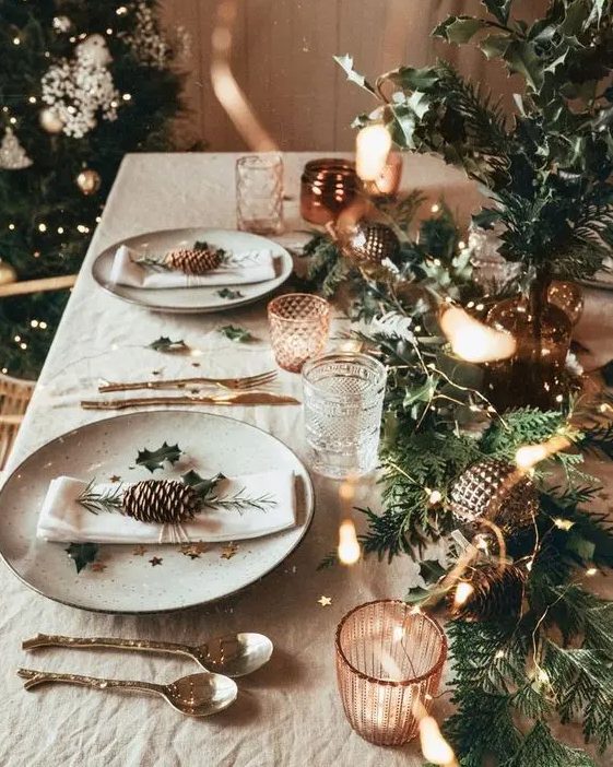 a bright christmas tablescape with an evergreen runner, lights, metallic ornaments, star print plates and copper candleholders