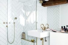 a bright glam white bathroom with chevron clad tiles, gilded touches, dark wooden beams and a square sink