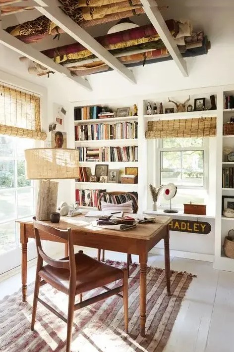 A catchy farmhouse home office with built in storage units and shelves, a wooden desk and chair, a rustic lamp and woven shades