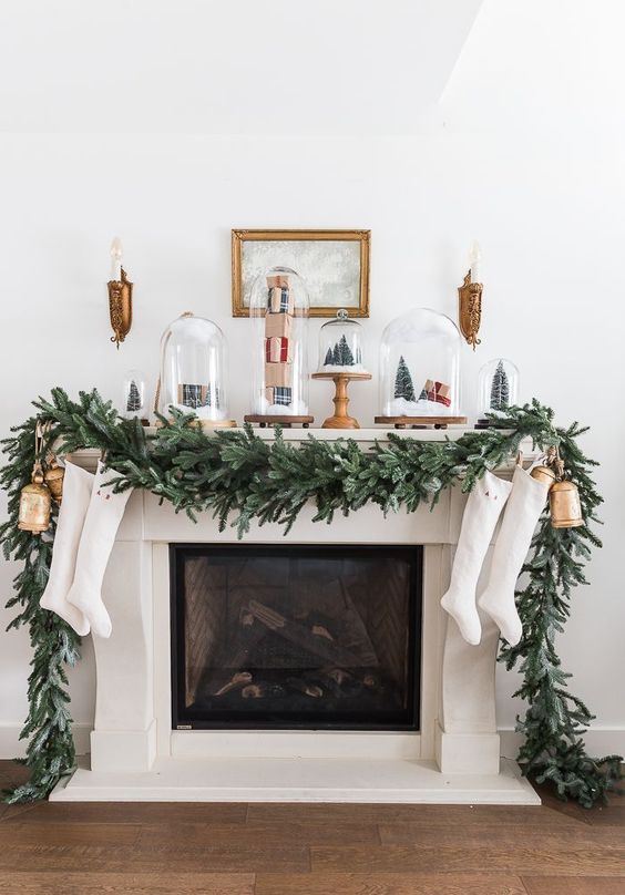 a chic Christmas mantel with a fir garland, white stockings, shiny gold bells, cloches with tinsel trees and gift boxes is amazing