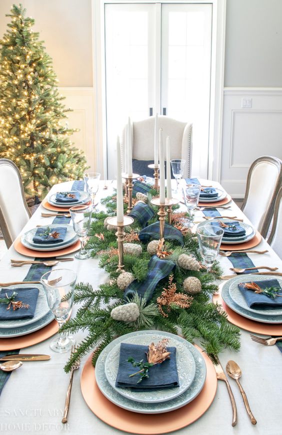 a chic Christmas tablescape with gold chargers and cutlery, blue plates and napkins, fir branches, glitter pinecones and candles