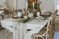 a chic Scandinavian Christmas table with gilded candleholders and lanterns, a neutral runner, greenery, gold bells over the table is amazing