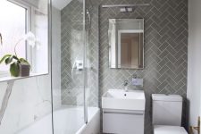 a chic modern bathroom with white marble and grey herringbone tiles, a tub with a glass screen, a chic chandelier