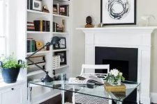 a chic modern neutral home office with built-in bookshelves, a fireplace, a glass desk, a neutral rug and a stylish chandelier