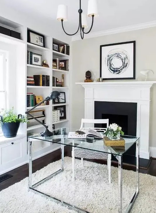 a chic modern neutral home office with built-in bookshelves, a fireplace, a glass desk, a neutral rug and a stylish chandelier