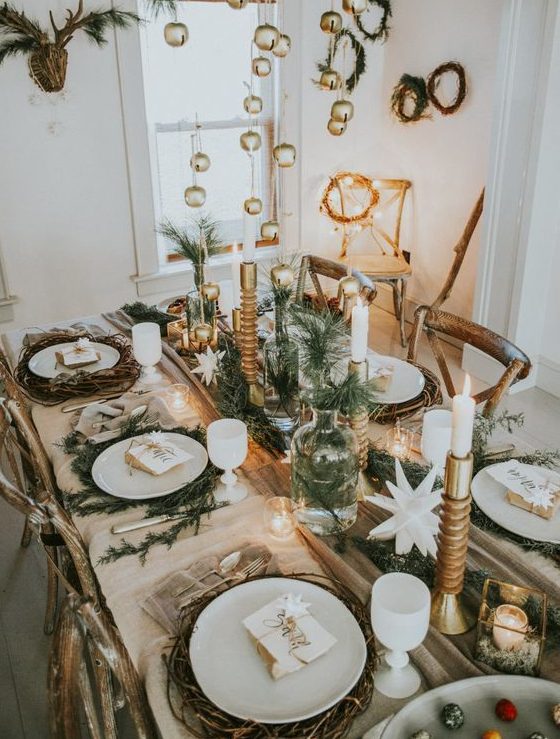 a chic rustic Christmas table with evergreens in bottles and vases, candles, woven placemats and bells hanging over the table
