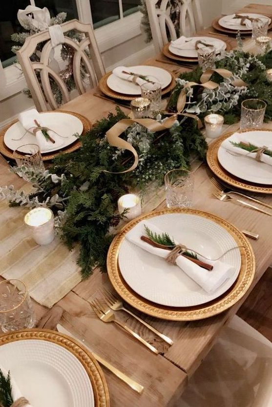 a chic rustic Christmas tablescape with gilded chargers, an evergreen runner with ribbons, pale branches, candles and cinnamon