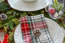 a cozy traditional table setting with evergreens, pinecones, plaid napkins and ornaments