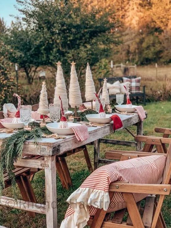 A creative outdoor Christmas tablescape with an evergreen runner, red and striped napkins, cone shaped trees and gnomes in plates