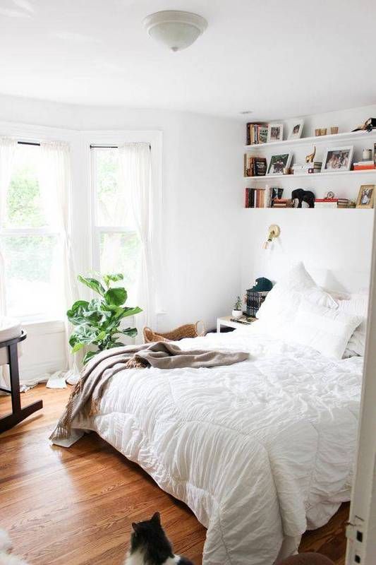 A light filled bedroom with a bow window, some shelves, a bed with neutral bedding, potted plants is welcoming