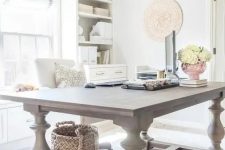 a light-filled farmhouse home office with a large wooden desk, neutral storage units, a crystal chandelier and neutral blooms