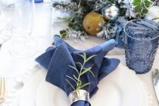 a lovely Christmas tablescape with navy linens, glasses, blooms and a candleholder, silver bells, white porcelain and chargers