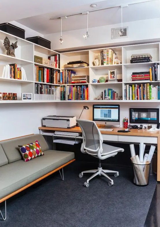 A mid century modern home office with a large open storage corner unit with lights, a desk and a white chair, a grey sofa