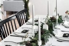 a minimalist Christmas tablescape with white linens, grey candles in white candleholders, an evergreen runner with pinecones and little houses
