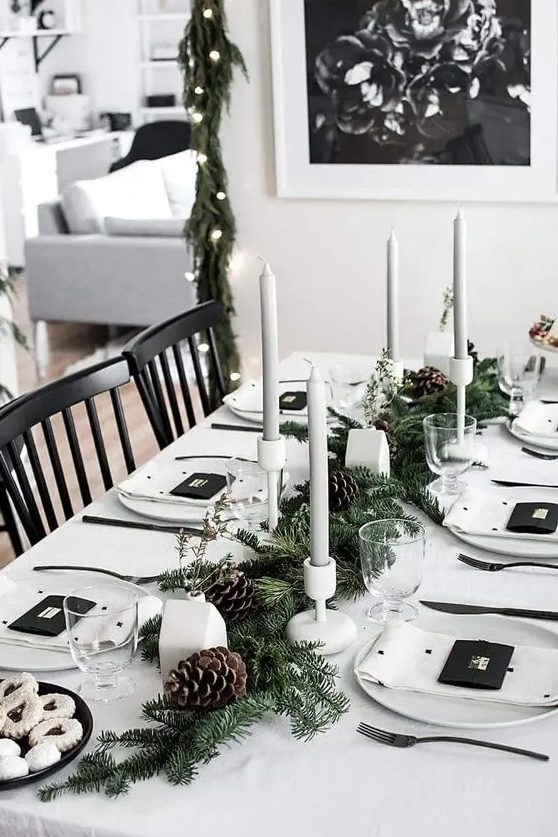 a minimalist Christmas tablescape with white linens, grey candles in white candleholders, an evergreen runner with pinecones and little houses