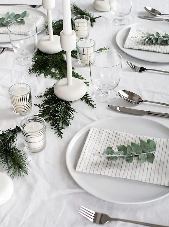 a minimalist table setting with an evergreen garland, white candles and eucalyptus for marking each place setting