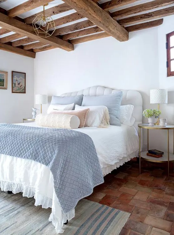 a modern farmhouse bedroom with wooden beams on the ceiling, a white bed with pastel blue and white bedding, tiered nightstands and a printed rug