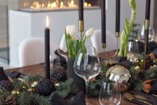 a modern moody Christmas table with gold chargers, blakc plates and napkins, black candles, white tulips and black ornaments and pinecones