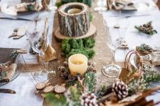 a natural Christmas table setting with a burlap runner, moss, tree stumps, snowy pinecones, tinsel trees and glasses