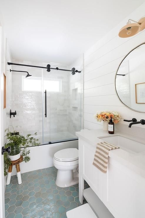 a neutral cottage bathroom with a bathtub and a shower space, a white vanity, a round mirror, a blue hexagon tile floor and a sconce