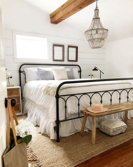 a neutral farmhouse bedroom with a metal bed, wooden nightstands and a bench, a crystal chandelier and neutral bedding
