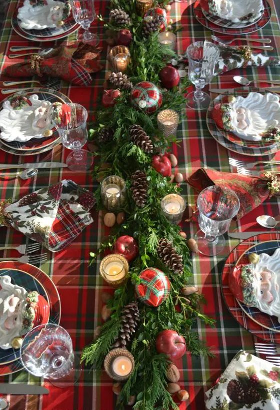 a plaid tablecloth, a greenery table runner with pinecones, apples, plaid ornaments and mercury glass candle holders