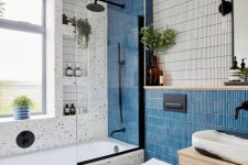 a pretty and cool bathroom with skinny blue and white tiles and terrazzo, a stained vanity, black fixtures and greenery
