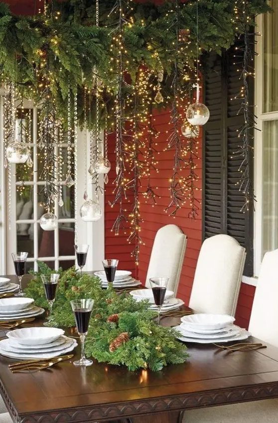 a pretty and elegant outdoor Christmas tablescape with an evergreen and pinecone runner with lights, white porcelain and lights over the table