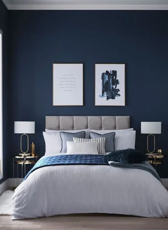 a refined modern bedroom with navy walls, a grey upholstered bed, chic artworks, gold nightstands and blue bedding