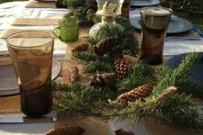 a rustic Christmas tablescape with a burlap table runner and placemats, clear glass chargers and evergreens, pinecones and table lamps