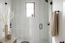 a serene neutral bathroom with white skinny and terrazzo tiles, a shower space with a window, a stained vanity, black fixtures