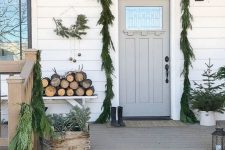 a simple and rustic Scandinavian Christmas porch with a fir garland, potted trees, a wreath with bells, firewood and crates with greenery