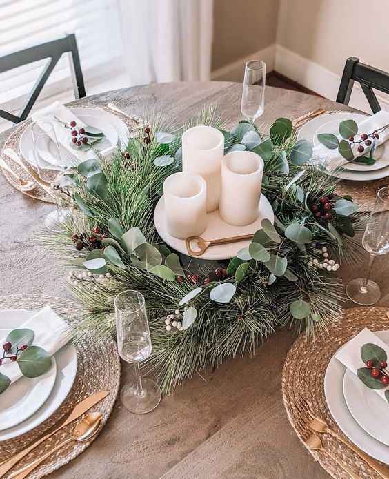 a simple yet glam Christmas tablescape with a greenery and berri wreath wrapping candles, woven chargers, gold cutlery and leaves