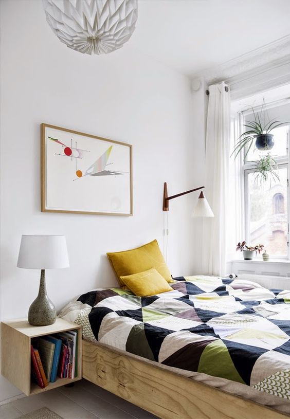 a small Scandinavian bedroom with a bed with bright bedding, a stained nightstand, a sconce and an artwork